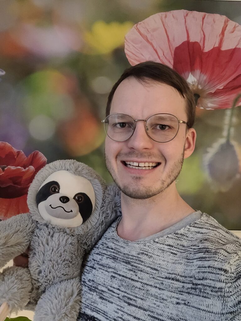 Das Bild zeigt Patrick Bannas, einen jungen Mann mit Brille und kurzen, blonden Haaren. Er hält ein Faultierkuscheltier im Arm. Im Hintergrund ist ein Bild mit Blumen zu sehen.